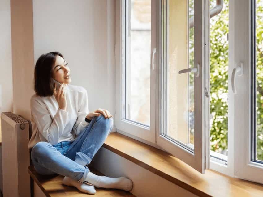uPVC Double Glazed Windows Options A woman sitting on a wooden window sill next to an open white uPVC casement window with a fixed central mullion.
